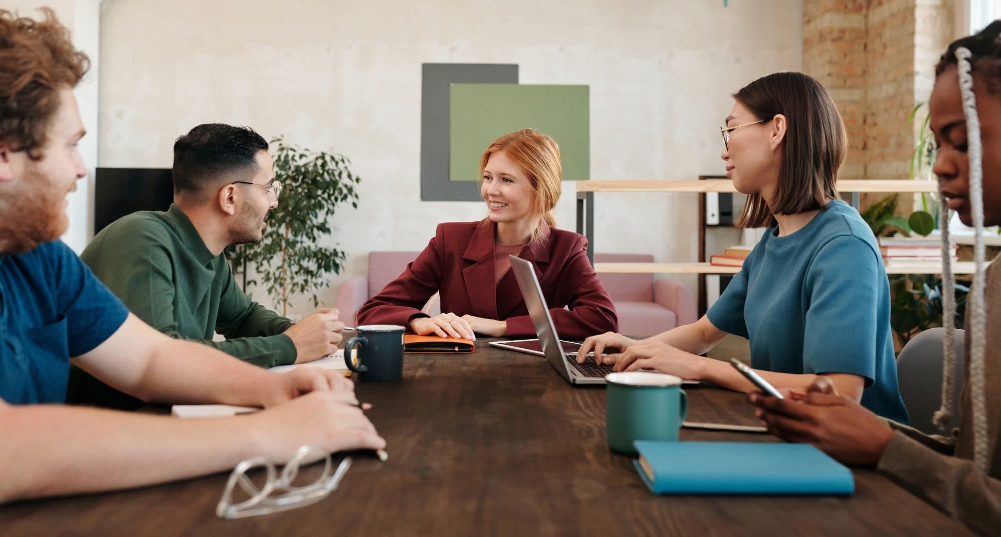Five people talking in a meeting around a wooden table, with laptops and coffee cups.